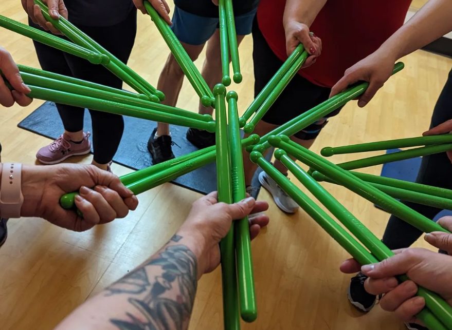 pound participants holding green drumsticks, called ripsticks, in a circle